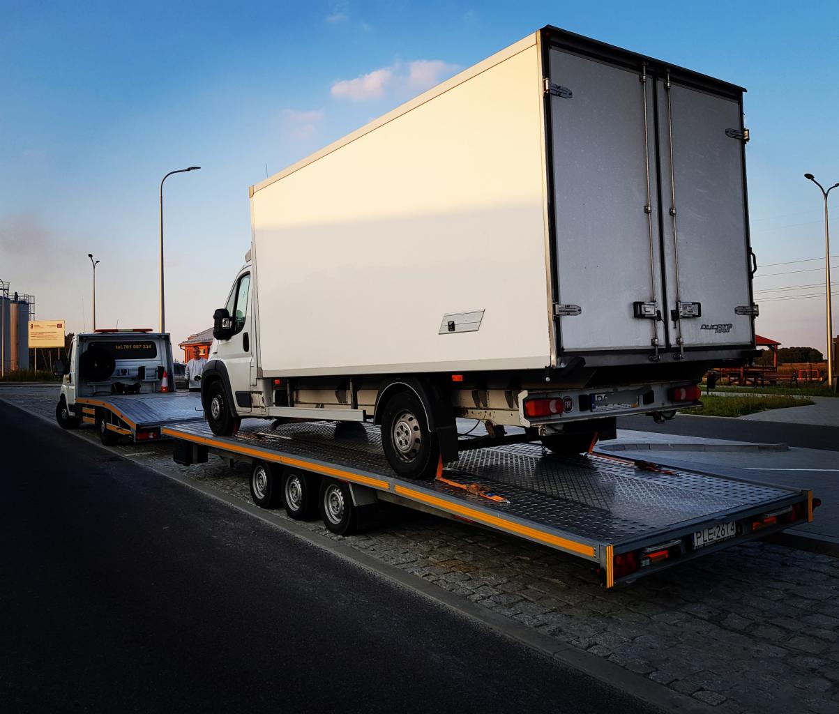 Biały samochód dostawczy zabezpieczony pomarańczowymi pasami na metalowej platformie lawety, ciągniętej przez mniejszy pojazd holowniczy na tle błękitnego nieba.