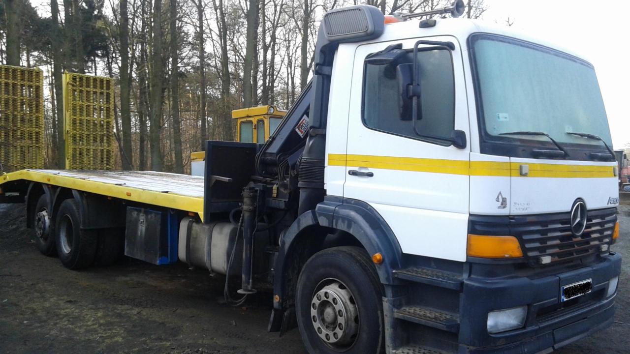 Żółto-biała laweta Mercedes-Benz z dźwigiem HDS na placu, widok z boku. Na platformie załadunkowej znajdują się ażurowe, żółte konstrukcje.