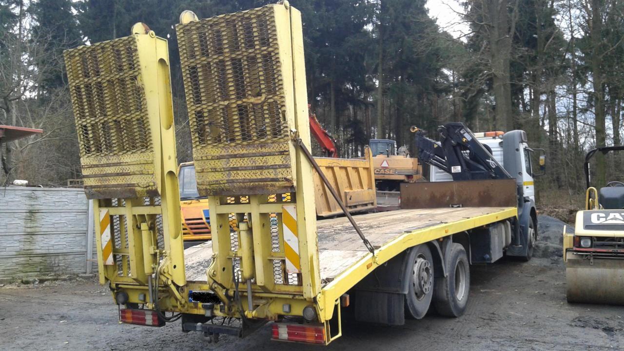 Żółta laweta z podniesionymi najazdami na tle drzew i innych maszyn budowlanych. Widoczny walec i ciężarówka z dźwigiem.