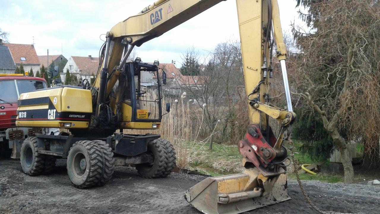 Żółta koparka kołowa CAT M318 na placu budowy, z widocznym łyżką i fragmentem czerwonej ciężarówki w tle, w otoczeniu drzew i zabudowań.