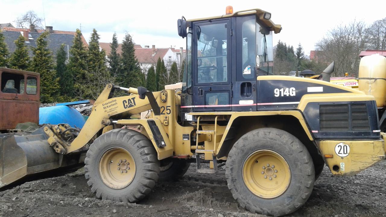 Żółta ładowarka CAT 914G z łyżką, widoczna z boku na placu budowy z ziemią, w tle drzewa i zabudowania. Maszyna gotowa do pracy.