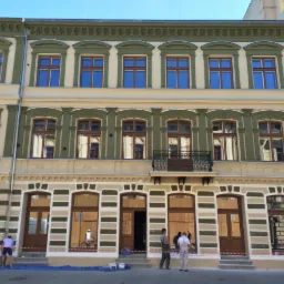 Odnowiona fasada kamienicy z zielonymi i kremowymi elementami dekoracyjnymi; ekipa remontowa pracuje na poziomie parteru, zabezpieczając okna i drzwi.