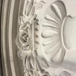 Zakład Budowlano-Sztukatorski - Detal dekoracyjny ściany z motywem róży, liści i wazy, wykończony białą farbą, widoczne nierówności powierzchni.