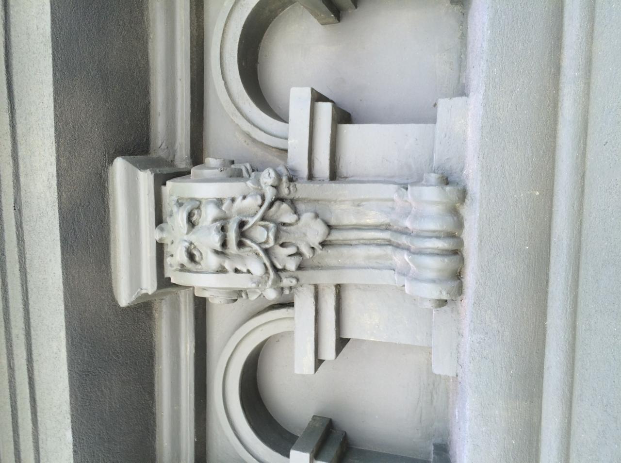 Detal architektoniczny w postaci płaskorzeźby z motywem roślinnym i geometrycznym, pomalowany na szaro, widoczny na tle ściany z wyraźną fakturą.
