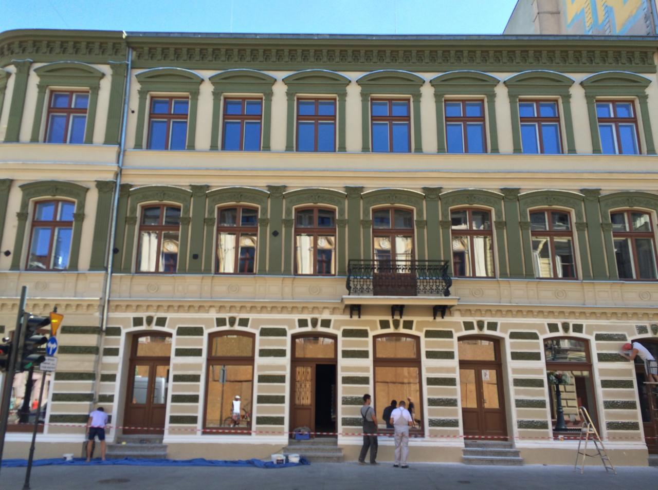 Odnowiona fasada kamienicy z zielonymi i kremowymi elementami dekoracyjnymi; ekipa remontowa pracuje na poziomie parteru, zabezpieczając okna i drzwi.