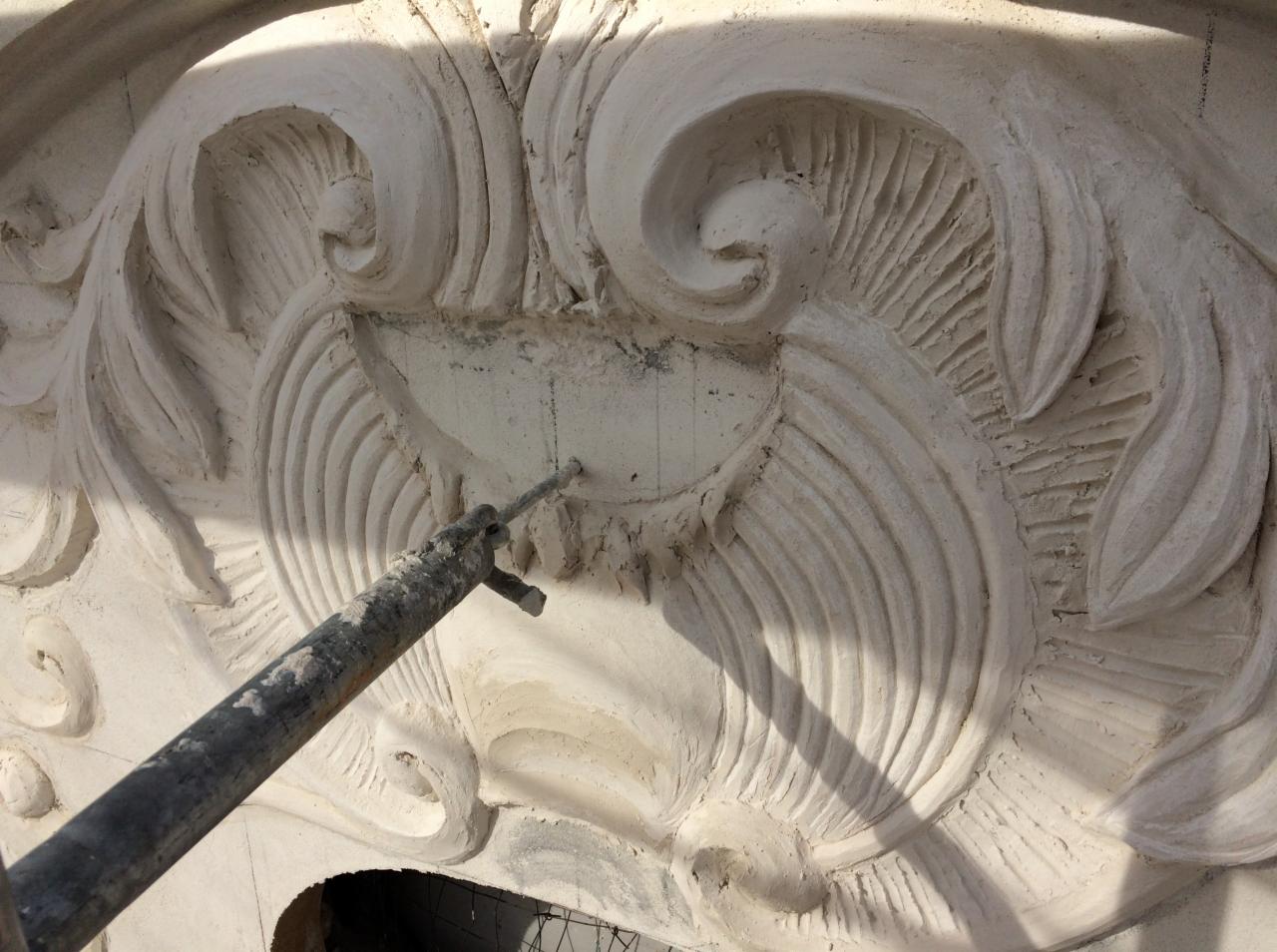 Dekoracyjny element architektoniczny z ornamentami w trakcie naprawy, widoczny fragment rusztowania i narzędzie wbite w powierzchnię.