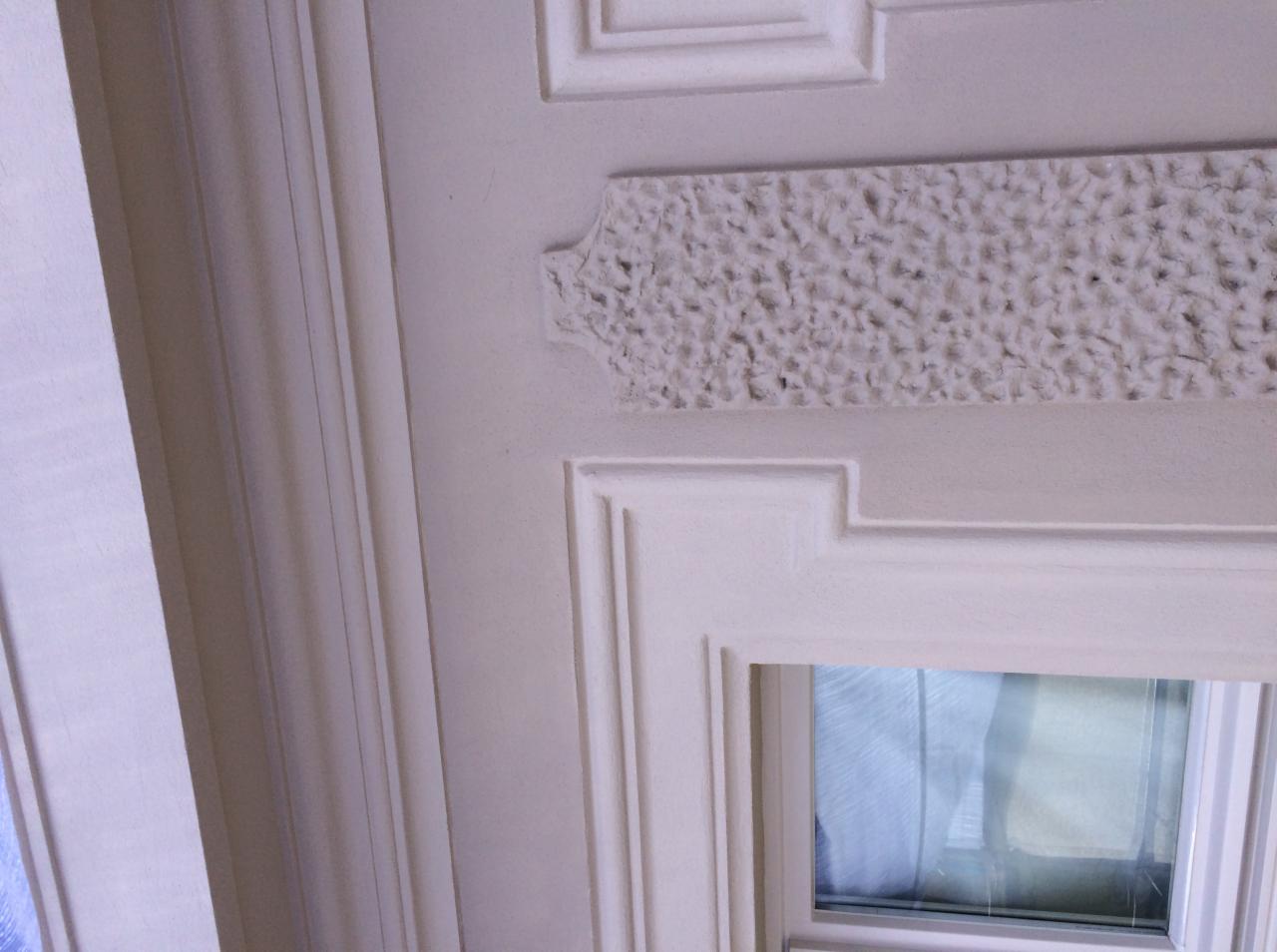 Fragment ściany z ozdobnym, teksturowanym pasem pod sufitem, pomalowanej na biało, z widocznym oknem w ozdobnej ramie.