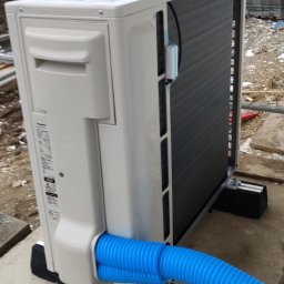 Evolui sp z o.o. - Zewnętrzna jednostka klimatyzacji zamontowana na betonowej podstawie, z podłączonymi dwoma niebieskimi, karbowanymi przewodami. Widoczne elementy montażowe i częściowo rusztowanie. W tle teren...