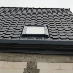 Ciemna dachówka ceramiczna z oknem dachowym w czarnej ramie, widok na fragment ściany z białych bloczków i szarego betonu, pochmurne niebo w tle.