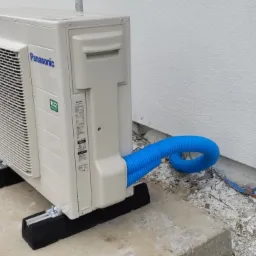 Jednostka zewnętrzna pompy ciepła Panasonic zamontowana na betonowej podstawie z niebieskim przewodem, widoczne ślady budowy i rusztowanie w tle.