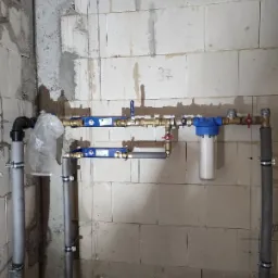 Szare rury wodne z niebieskimi elementami instalacji zamontowane na ścianie z białych bloczków, widoczny filtr wody z białym wkładem.