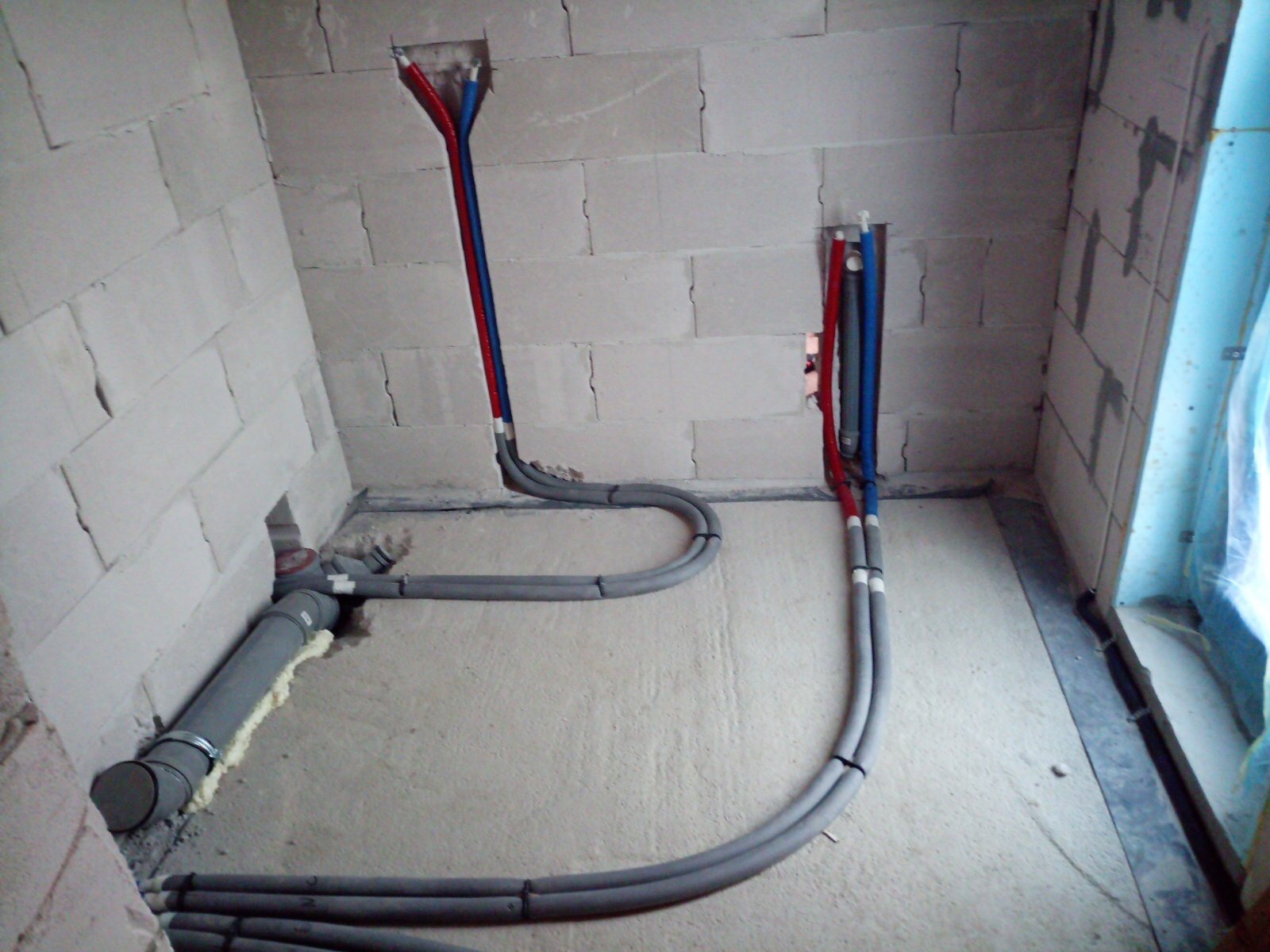 Szare rury kanalizacyjne i pionowe rury wodne (czerwona i niebieska) w surowym wnętrzu z białych bloczków, widoczne mocowania i pianka montażowa.