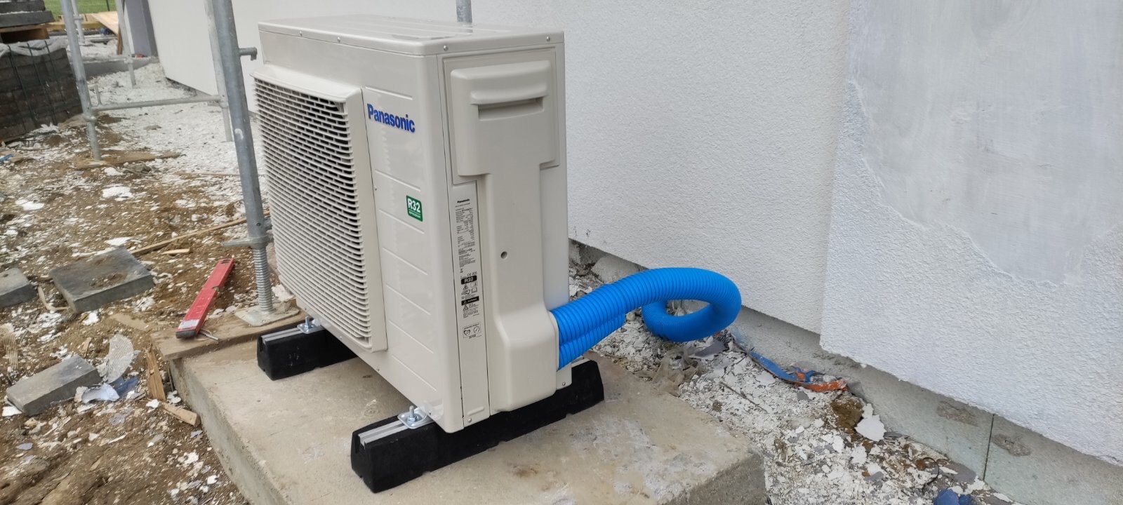 Zewnętrzna jednostka pompy ciepła Panasonic zamontowana na betonowej podstawie z niebieskim karbowanym przewodem odprowadzającym skropliny przy ścianie budynku w trakcie budowy.
