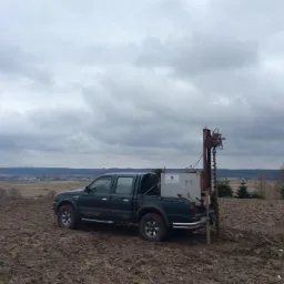 Ciemnozielony pickup z wiertnicą na pace, wykonujący odwiert w polu, widok na odległy krajobraz pod pochmurnym niebem.