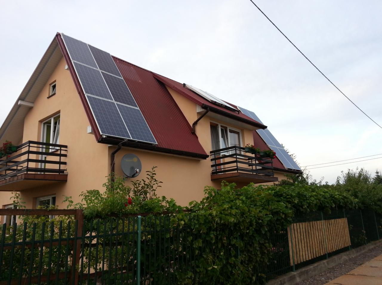 Dom jednorodzinny z czerwonym dachem, na którym zamontowano panele słoneczne; widoczna antena satelitarna na ścianie i balkon z kwiatami.