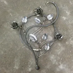 Kuty element dekoracyjny w kształcie róży z liśćmi, wykonany z metalu, na szarym tle. Detal ogrodzenia lub balustrady, prezentujący kunszt kowalski.