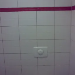 Wykończona łazienka z białymi płytkami i bordowym pasem dekoracyjnym, widoczny przycisk spłuczki na ścianie.