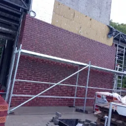 Wznoszenie ściany z cegły klinkierowej z widocznym ociepleniem wełną mineralną na zewnętrznej ścianie budynku, z użyciem rusztowania i palet z cegłami.