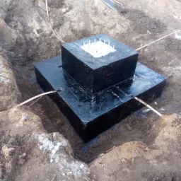 Fundament pod dom w wykopie, zabezpieczony czarną, błyszczącą izolacją, z widocznymi prętami zbrojeniowymi zalane betonem, na ziemi leżą kable uziemiające.