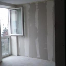 PRZEDSIĘBIORSTWO USŁUGOWO - HANDLOWE SAN-MARC FILIP MARCHELA - Wnętrze pokoju w trakcie wykańczania. Widoczne ściany z zamontowanymi płytami karton-gips, częściowo zaszpachlowane łączenia. Okno balkonowe z widokiem na zewnątrz.