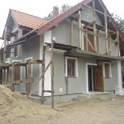 PRZEDSIĘBIORSTWO USŁUGOWO - HANDLOWE SAN-MARC FILIP MARCHELA - Dom w trakcie prac elewacyjnych: szary tynk, drewniane rusztowania, brązowa stolarka okienna i drzwiowa, sterta piasku przed budynkiem. Częściowo ocieplony.