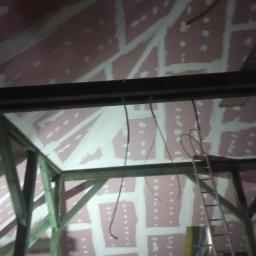 PRZEDSIĘBIORSTWO USŁUGOWO - HANDLOWE SAN-MARC FILIP MARCHELA - Poddasze w trakcie prac wykończeniowych: widoczne drewniane elementy konstrukcyjne, różowe płyty z wypełnionymi spoinami oraz zwisające przewody elektryczne i drabina oparta o konstrukcję.