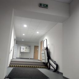 PRZEDSIĘBIORSTWO USŁUGOWO - HANDLOWE SAN-MARC FILIP MARCHELA - Wnętrze korytarza z windą schodową dla osób niepełnosprawnych, widoczne schody z oznaczeniem ostrzegawczym, tablica informacyjna i znak ewakuacyjny.