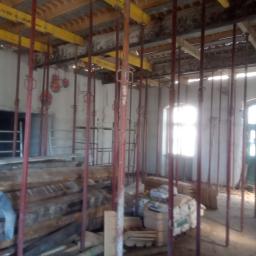 PRZEDSIĘBIORSTWO USŁUGOWO - HANDLOWE SAN-MARC FILIP MARCHELA - Wnętrze budynku w trakcie budowy z widocznymi podporami stropowymi w kolorze czerwonym i żółtym, stertą drewna i materiałów budowlanych, oraz oknem w tle.