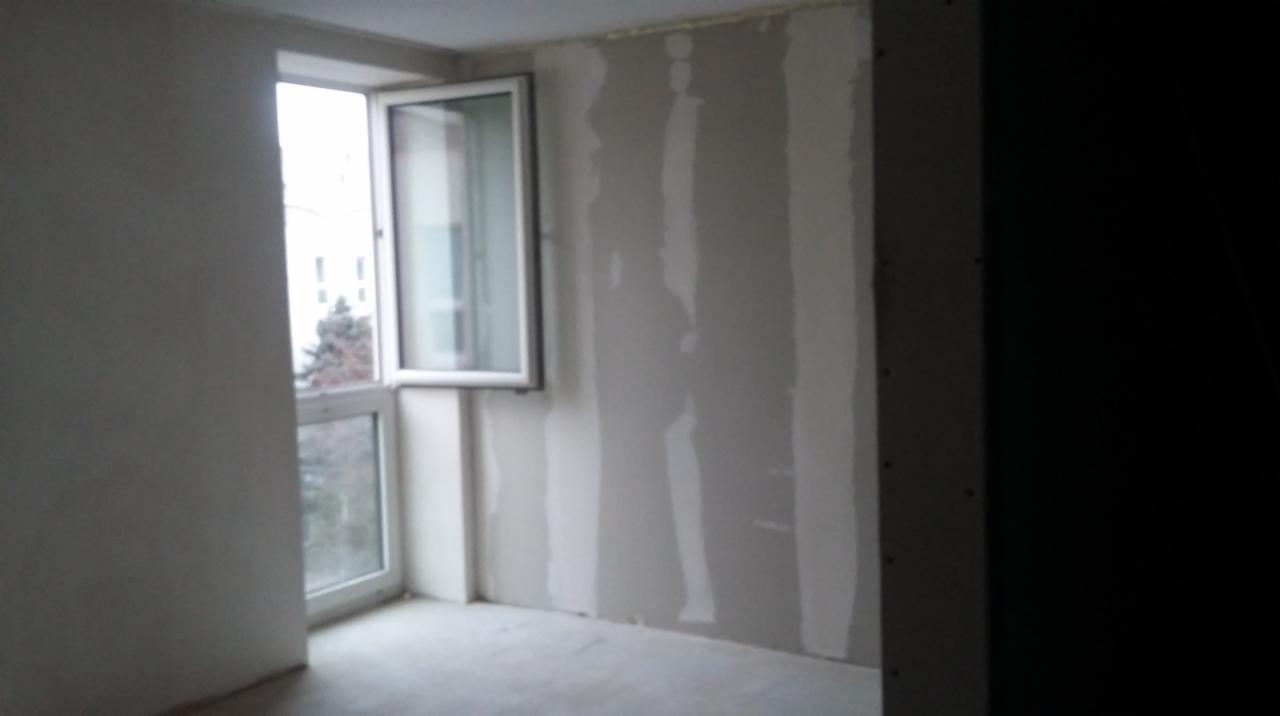 Wnętrze pokoju w trakcie wykańczania. Widoczne ściany z zamontowanymi płytami karton-gips, częściowo zaszpachlowane łączenia. Okno balkonowe z widokiem na zewnątrz.
