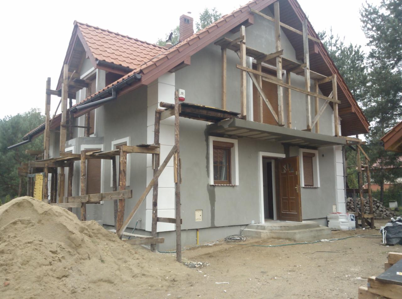 Dom w trakcie prac elewacyjnych: szary tynk, drewniane rusztowania, brązowa stolarka okienna i drzwiowa, sterta piasku przed budynkiem. Częściowo ocieplony.