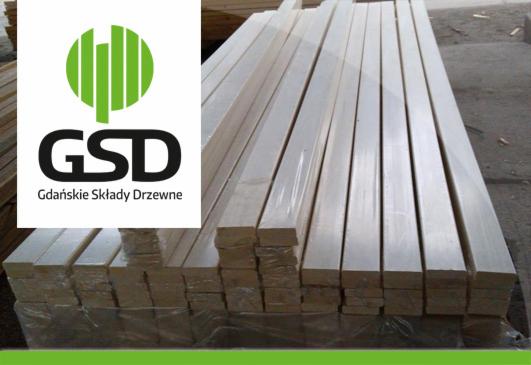 Stos tarcicy budowlanej owiniętej folią, z logo Gdańskie Składy Drzewne w lewym górnym rogu. Widoczne jasne drewno i ciemne tło magazynu.