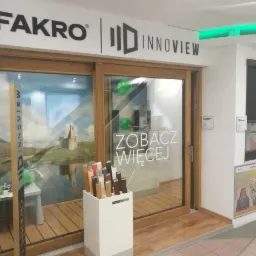 Ekspozycja okien i drzwi przesuwnych w salonie firmowym Fakro Innoview, z widocznymi próbkami kolorów i faktur drewna oraz roletą zewnętrzną.
