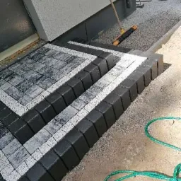 Schody wejściowe wyłożone kostką brukową w odcieniach szarości i czerni, z białymi akcentami, zakończone obrzeżem z kostki. W tle widoczna ściana budynku i częściowo uprzątnięty teren z resztkami...