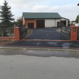 KUL-BRUK - Ukończony podjazd z czarnej kostki brukowej z obramowaniem z czerwonej kostki, prowadzący do garażu z zielonym dachem, widoczna brama z kutego żelaza i słupki z cegły, na jednym ze słupków skrzynka...