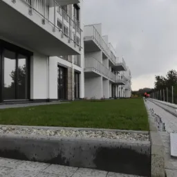 Nowoczesny budynek apartamentowy z balkonami, świeżo założony trawnik z obrzeżem z kamienia i kostki brukowej, widoczny fragment chodnika z odwodnieniem liniowym w trakcie montażu.