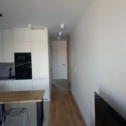 Wnętrze mieszkania po remoncie: widok na kuchnię z białymi szafkami, drewniany blat stołu z przezroczystym krzesłem, parkiet, białe ściany i drzwi w głębi korytarza.