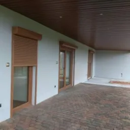 Zewnętrzny taras z brązową kostką brukową, na ścianie okna i drzwi z drewnianą ramą, zabezpieczone zwiniętymi roletami w kolorze drewna, pod drewnianym zadaszeniem.