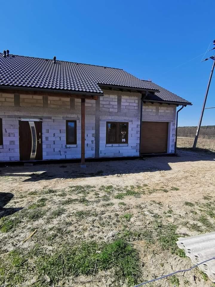 Surowy stan budynku z szarego bloczka z zamontowanymi brązowymi drzwiami wejściowymi i bramą garażową, widoczne okna z brązowymi ramami, ciemna dachówka na dachu, słup energetyczny w tle.