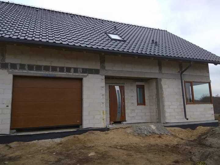 Surowy stan domu jednorodzinnego z brązową segmentową bramą garażową i oknami z brązowymi ramami, widoczne niedokończone elewacje i teren po budowie.