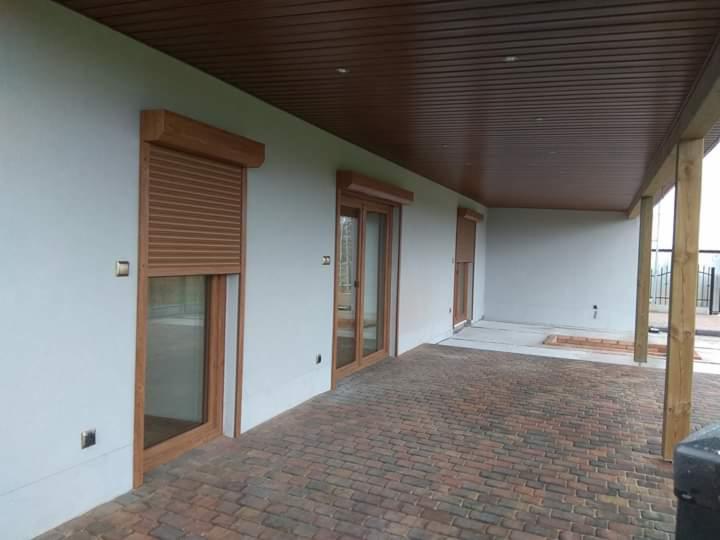 Zewnętrzny taras z brązową kostką brukową, na ścianie okna i drzwi z drewnianą ramą, zabezpieczone zwiniętymi roletami w kolorze drewna, pod drewnianym zadaszeniem.