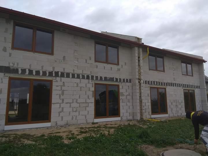 Surowy stan budynku z brązowymi oknami i drzwiami przesuwnymi, widoczne ślady zaprawy, trawa na pierwszym planie, pochmurne niebo.