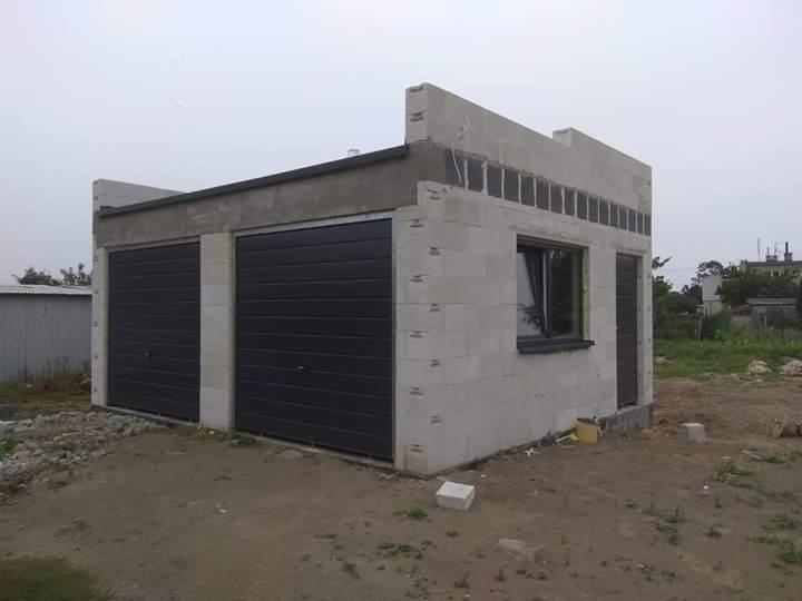 Surowy garaż z dwoma ciemnymi bramami segmentowymi, oknem i drzwiami, w trakcie budowy z bloczków betonowych, na niezagospodarowanym terenie.