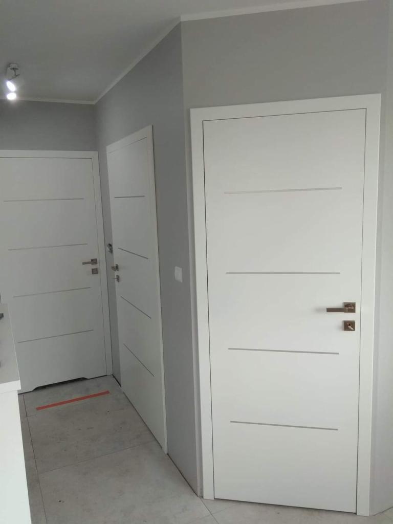 Trzy białe, nowoczesne drzwi wewnętrzne z poziomymi frezowaniami i klamkami w odcieniu miedzianym, ustawione w rzędzie w szarej ścianie.