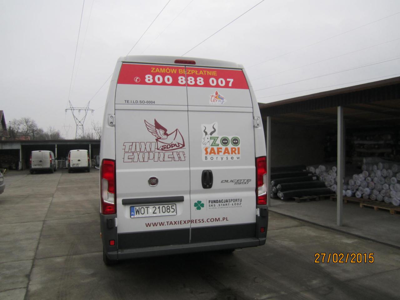 Tył białego busa marki Fiat Ducato z reklamami Taxi Express, Zoo Safari Borysew i numerem telefonu zamówień, zaparkowanego na betonowym placu w pochmurny dzień.