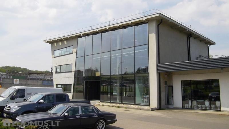 Nowoczesny budynek biurowy z fasadą ze szkła strukturalnego, zaparkowane samochody i busy, widok z zewnątrz, szare niebo.