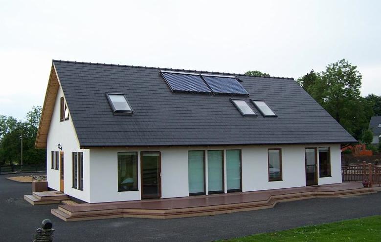 Biały dom z ciemnym dachem, panelami słonecznymi i oknami dachowymi. Widoczny taras i podjazd. W tle drzewa i maszyna budowlana.
