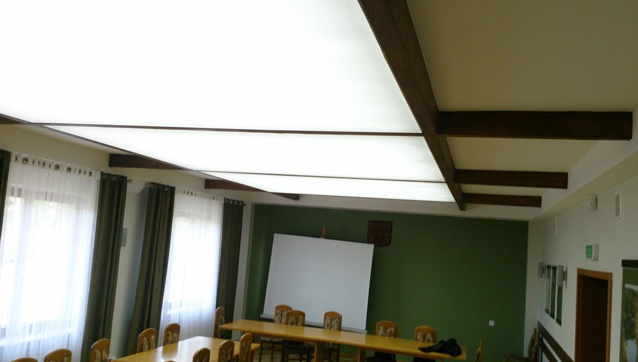 Sala konferencyjna z podwieszanym sufitem napinanym i drewnianymi belkami, oświetlonymi od góry. Widoczne okna z zasłonami i długi stół.