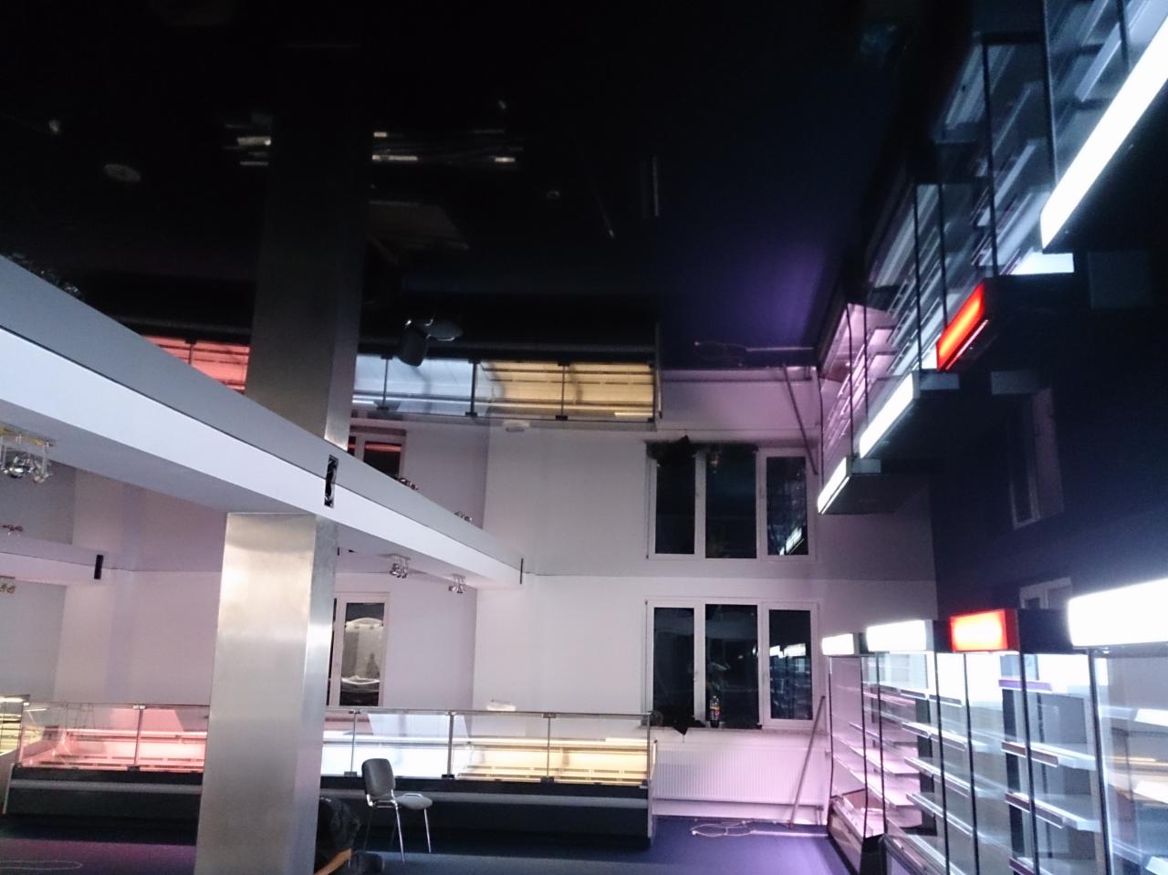 Wnętrze lokalu usługowego w trakcie adaptacji: widoczne metalowe słupy, instalacje pod sufitem, okna i regały. Lokal w stanie surowym, przygotowywany do użytku.