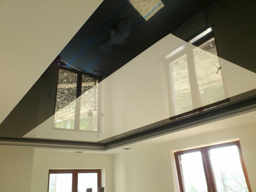 Biały, błyszczący sufit napinany z ciemną obwódką, odbijający okna. Widoczny fragment instalacji nad sufitem. Ujęcie z dołu, w jasnym wnętrzu z oknami.