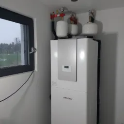 Biała pompa ciepła marki Stiebel Eltron z trzema białymi zbiornikami wyrównawczymi i czerwonymi elementami instalacji, zamontowana w jasnym pomieszczeniu z oknem, przez które widać zimowy krajobraz.